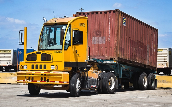 Yellow Kalmar container chassis truck with red shipping container on gravel yard with other industrial vehicles